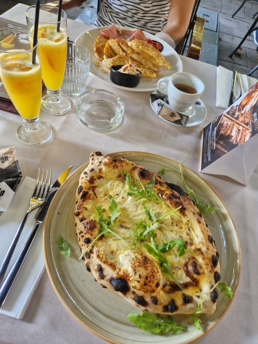 A restaurant table features a large pizza topped with arugula, along with glasses of orange juice, a coffee cup, and a plate of breakfast items.