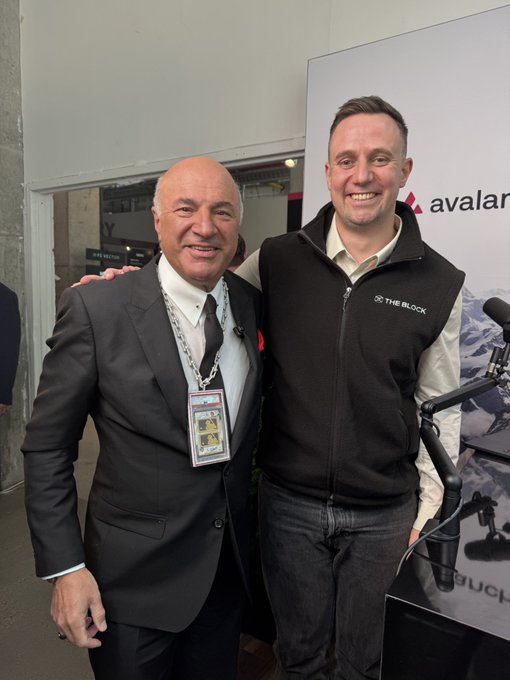 Two men pose together at an event, with one wearing a suit and lanyard and the other in a 'The Block' vest, standing in front of an Avalanche logo backdrop.