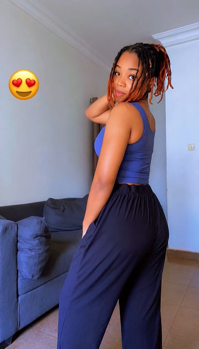 A photo of a young woman with reddish-brown locs wearing a purple crop top and dark navy pants, posing in a living room with a gray sofa, with a heart-eyes emoji overlay in the upper left corner.