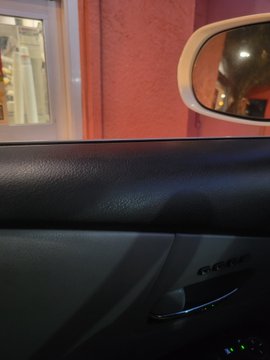 In this blurry scene, a car interior is visible with a grey car door next to a window and an open door in the background. A person is sitting inside another car, which appears to be a minivan. The driver's side mirror is reflected through the window, suggesting that the car has been used recently or was parked near a reflective surface. There are also other cars visible outside of the frame, but they are not prominently featured in the scene.