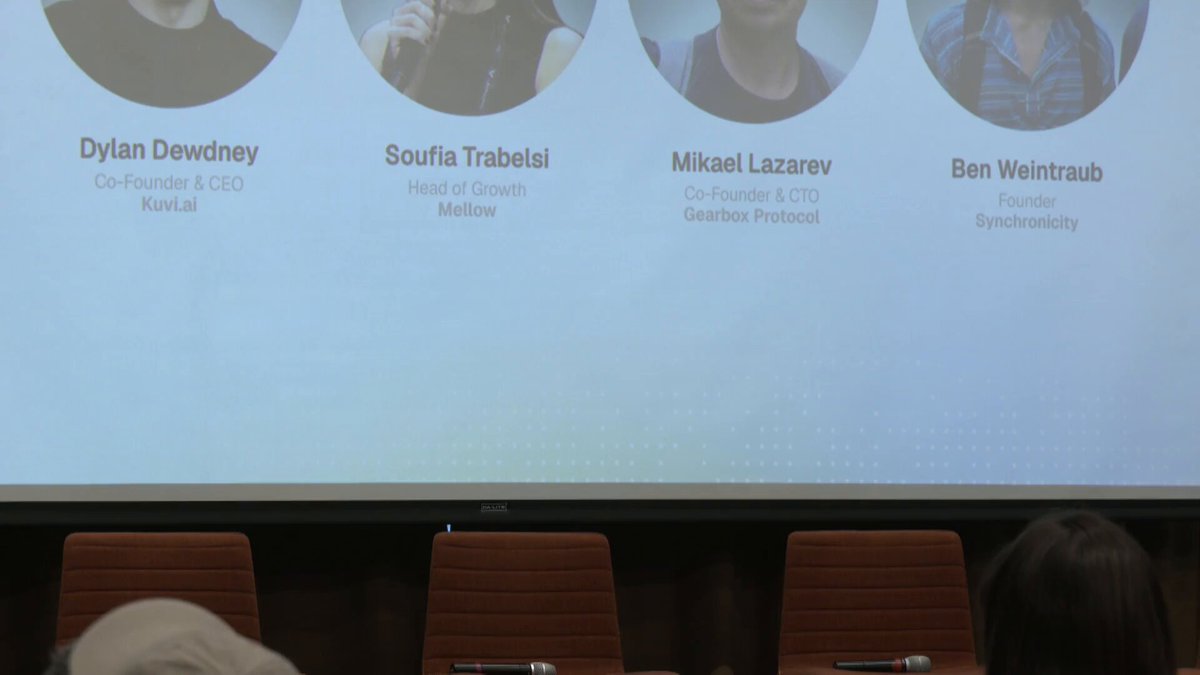 The image features a large projector screen with information about four people sitting behind their respective chairs, looking at the screen as if they are participating or observing an event. The panel includes individuals from various backgrounds such as Dylan Dewdney, Soufia Trabelsi, Mikael Lazarev, and Ben Weintraub. They seem to be engaged in a discussion about a particular topic of interest.