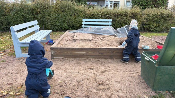 Two young children in dark blue winter jackets play in a sandy playground area, with one playing in a wooden sandbox and another standing nearby holding a green toy.