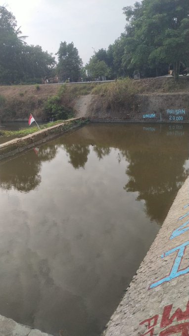 A photo shows a murky water reservoir or canal with text painted on the concrete reading 'BRISAN 2.000' and other markings, with a small red and white flag on the edge.