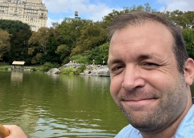 A selfie of a smiling middle-aged man with short hair in front of a pond in Central Park, New York, with buildings and trees visible in the background.