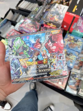 A hand holds a "Dragon Ball Super Card Game: Ultimate Booster" box featuring Frieza, with visible "DB-04" packaging, while stacks of other booster boxes fill the background of the TCG show. The scene aligns with the post's description of an active trading card event in Dubai, though the cards being showcased appear to be Dragon Ball Super TCG rather than VeeFriends.