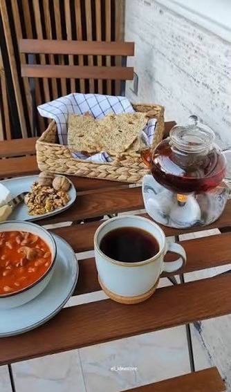 A table setting featuring a cup of dark tea or coffee, a bowl of soup, flatbread in a basket, and a glass teapot.