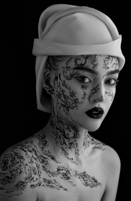 A black and white artistic portrait of a woman wearing a white head covering, with intricate tattoo-style patterns covering her face, neck, and shoulders, featuring dark lipstick.