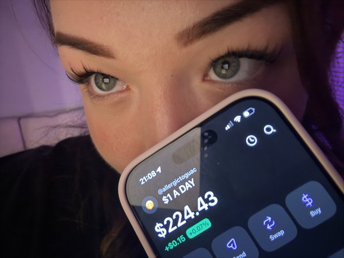 A close-up of a person with green eyes holding a smartphone displaying the $A DAY token on what appears to be a crypto wallet app, showing a value of $224.43 (+3.07%).