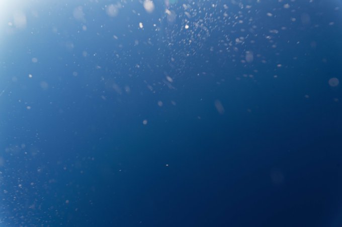 Sunlight filters down from the surface through deep blue water, illuminating scattered particles and bubbles that float suspended in the ocean depths. The image embodies the color "Blue" through its rich, gradient hues ranging from bright cerulean near the surface to dark navy in the abyss below.