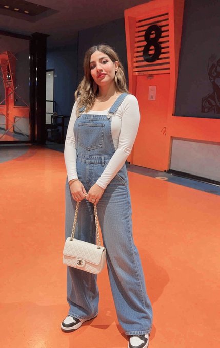 A young woman with long blonde hair poses in a casual outfit featuring blue denim overalls over a cream long-sleeve top, holding a white quilted chain-strap handbag, with a large "8" visible on the red wall behind her. The vibrant, stylish photo complements the cheerful morning greeting, reflecting a pretty and upbeat start to the day.