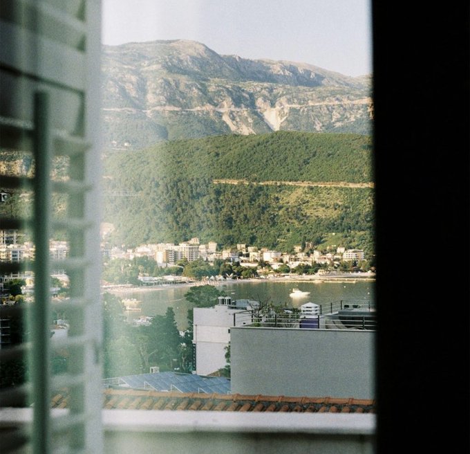 A scenic view through a window reveals a coastal town nestled between lush green hills and a bay with boats, with mountains rising in the background. The peaceful morning vista contrasts with the caption's enthusiastic message about memestocks and crypto innovation at Token2049, suggesting a moment of reflection amid the fast-paced world of blockchain developments.