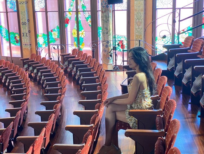 A woman with long dark hair in a floral dress sits alone in the wooden pews of an empty auditorium, with colorful stained glass windows illuminating the scene behind her.