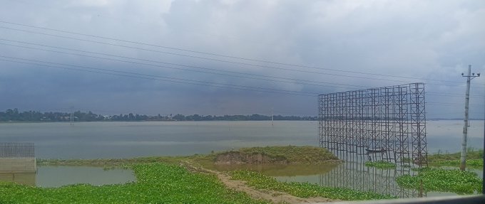 A landscape view of a large body of water under a cloudy sky, with a metal billboard frame, power lines, and green vegetation in the foreground.