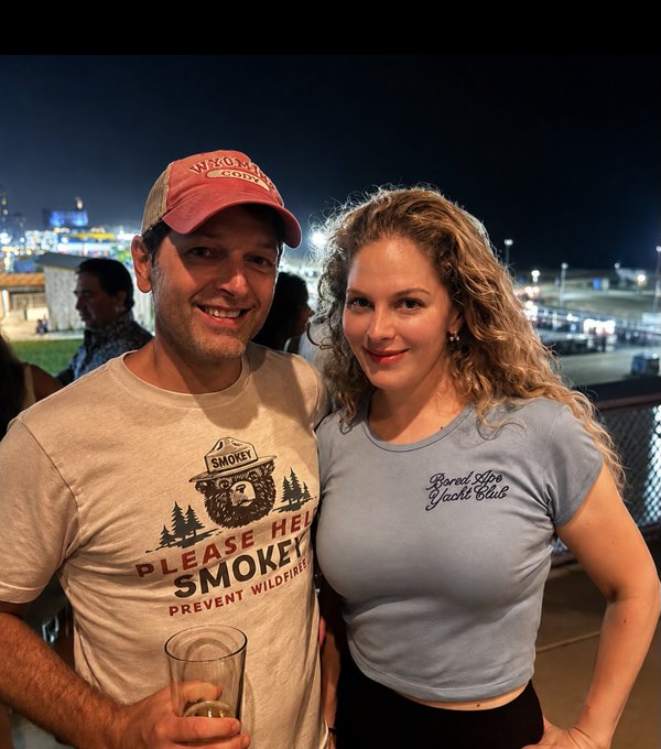 A man wearing a 'Please Help Smokey Prevent Wildfires' t-shirt and a baseball cap poses with a smiling woman wearing a 'Barrel Life Yacht Club' t-shirt at what appears to be an outdoor nighttime event.