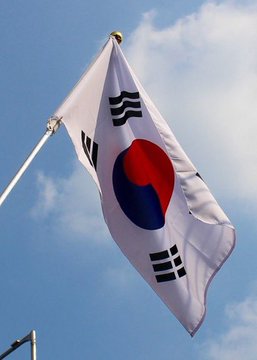 South Korean flag (Taegeukgi) waving on a flagpole against a blue sky with white clouds, showing the red and blue taegeuk symbol and black trigrams.