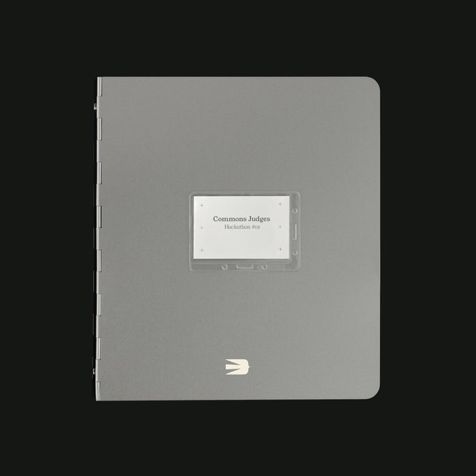 A gray notebook or binder with metal rings sits on a black background, featuring a clear card holder with a label reading 'Commons Judges Hackathon #02' and a small bird logo.