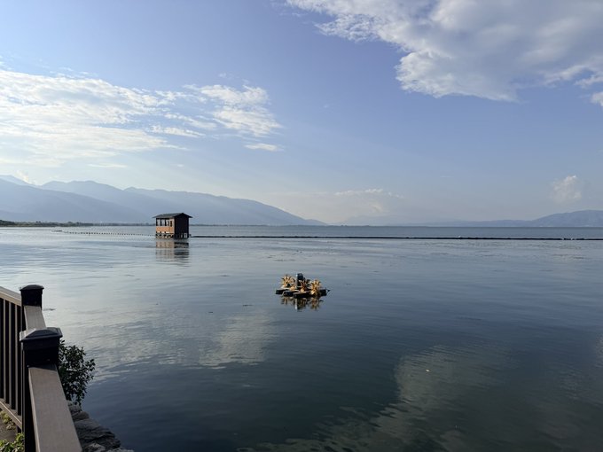 A serene lake scene with a small boat carrying passengers in yellow life vests, a wooden structure on stilts extending across the water, and mountains in the hazy distance under a blue sky.