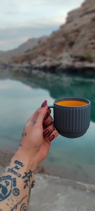 A tattooed hand with dark red nail polish holds a gray mug of orange liquid, with a serene lake and rocky cliffs in the background.