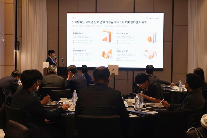 The image shows a conference or presentation setting where attendees are seated at round tables facing a large presentation screen displaying Korean text about digital asset services and customer experience.