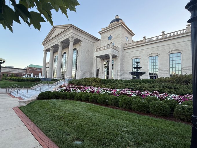 An exterior view of a grand classical-style building with tall columns, a clock tower, and a tiered fountain in the foreground, surrounded by landscaped gardens with colorful flowers and green lawns.
