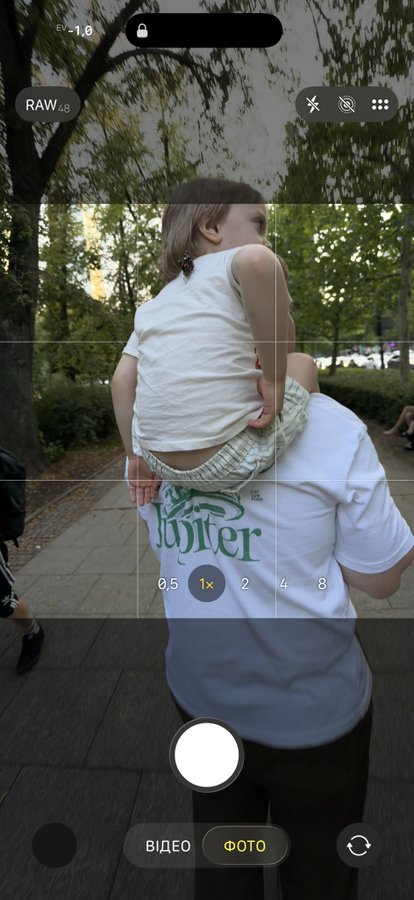 The image shows a camera app viewfinder with a child riding on an adult's shoulders in a park setting, zoom set at "1x" with "RAW 48" and exposure "-1.0" visible on screen. The photo humorously illustrates the "Active Shoulder Rewards" caption by capturing a literal child perched on someone's shoulders.