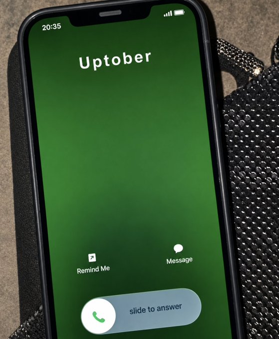 A smartphone displaying an incoming call screen with the caller ID 'Uptober' on a green background, set against a black beaded bag.