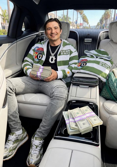 A man sitting in the back of a luxury car holding a large stack of cash, wearing a green and white striped sweater with dollar sign patches, a chain necklace, and designer sneakers, with more cash and merchandise visible in the seat beside him.