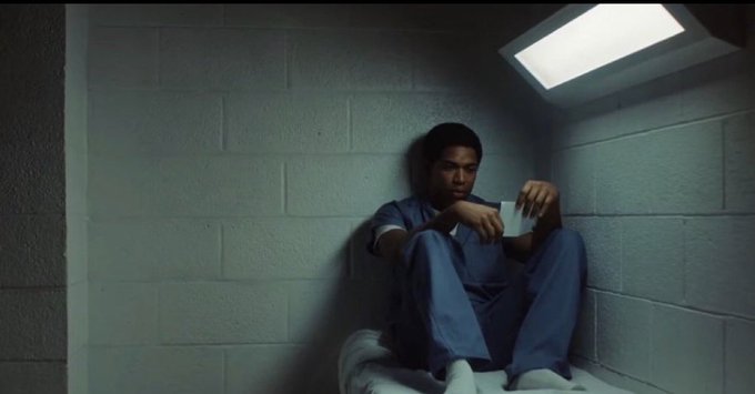 A man in blue clothing sitting on a bed in a stark, dimly lit prison room, holding a small cup.