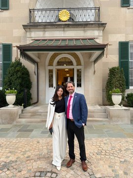 A man in a blue suit and pink tie poses with a woman in a black and white outfit in front of an elegant building entrance.