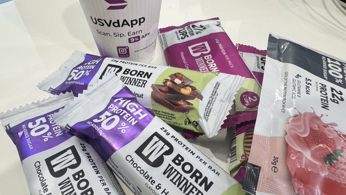 Several packages of Born Winner high protein bars in various flavors including Chocolate & Hazelnut are scattered on a surface alongside a UsvdApp branded cup.