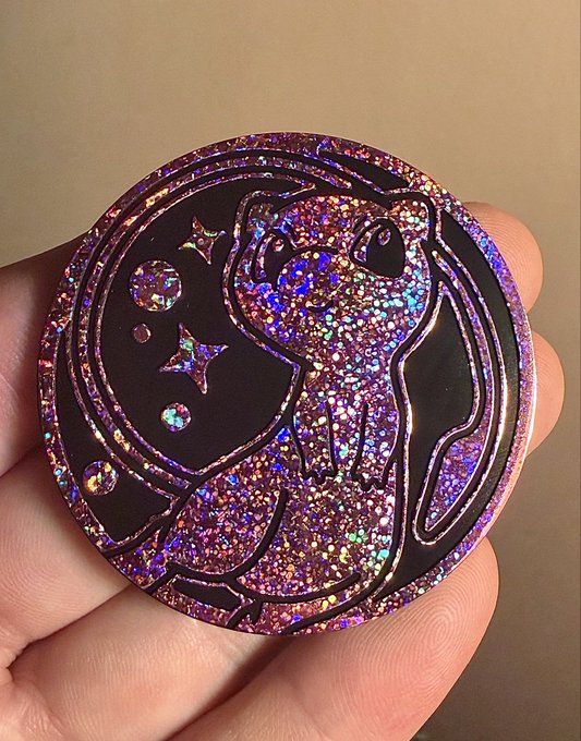 A close-up photo of a glittery purple and pink enamel pin featuring an animal design with stars and planets, held in someone's hand.