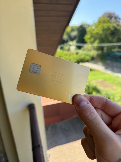 A hand holds a gold-colored crypto debit card with a smart chip outdoors, with a green landscape and sky visible in the background.