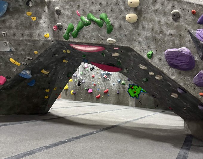 An indoor rock climbing gym featuring a distinctive curved/arched climbing wall structure with colorful holds in various shapes and colors.
