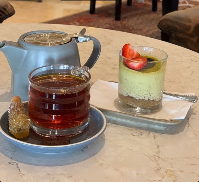 A blue teapot sits beside a glass cup of dark tea with a sugar-coated treat and a dessert glass layered with cream, crumbs, and fresh strawberries on a marble table.
