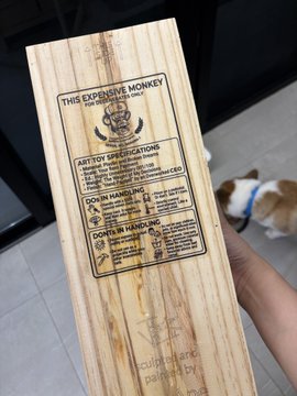 A wooden plaque labeled 'THIS EXPENSIVE MONKEY' being held up, with sections titled 'ART TOY SPECIFICATIONS', 'DOESN'T HANDLE', and 'DOES HANDLE', with a small dog visible in the background.