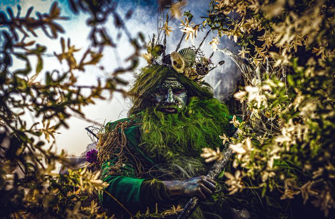 A fantasy figure with a green mossy beard, decorative face paint, and a headdress adorned with feathers and flowers emerges from a forest of foliage.