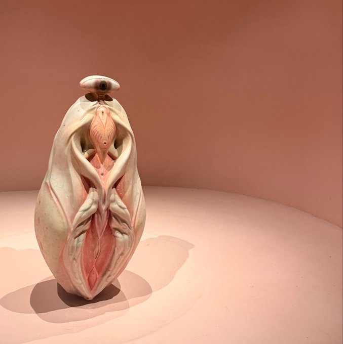 A surreal ceramic sculpture with an organic, biomorphic shape featuring pink and cream coloring, displayed on a pink surface.