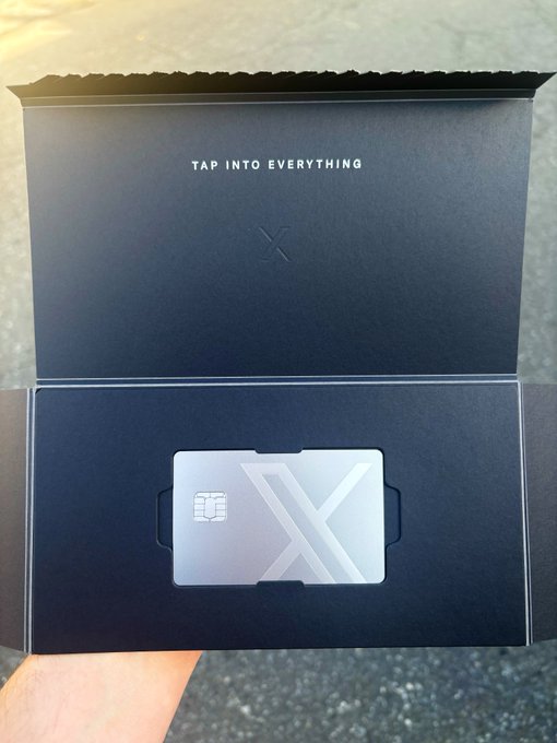 A metallic X-branded metal credit card inside a black premium packaging box with the text 'TAP INTO EVERYTHING', held by a hand.
