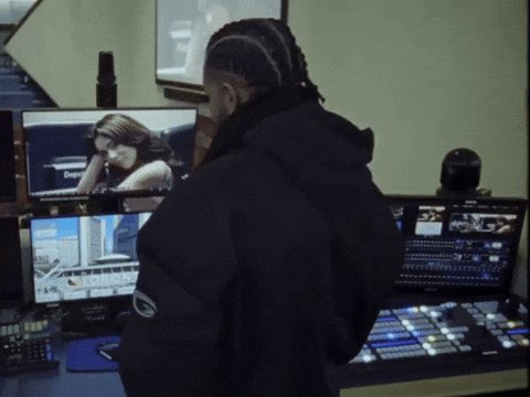 The image shows a man wearing a black hoodie standing behind a desk with two computer monitors, one of which displays an image of a woman's face. The man appears to be looking at the monitor or possibly working on a project related to the image. There is also a microphone and a TV in the scene.