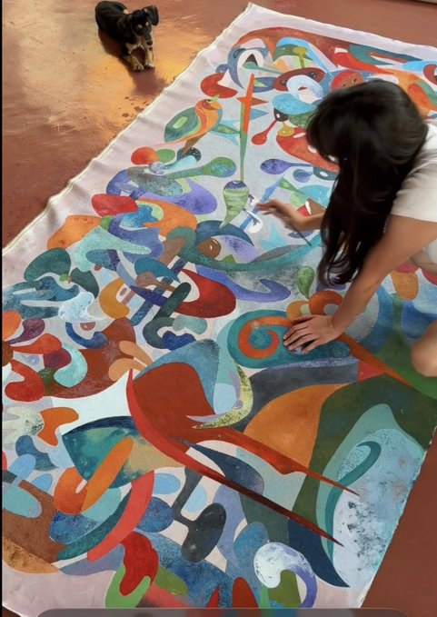 A person kneeling on a floor painting a large, colorful abstract rug with swirling patterns, while a small dog watches from the wooden floor nearby.