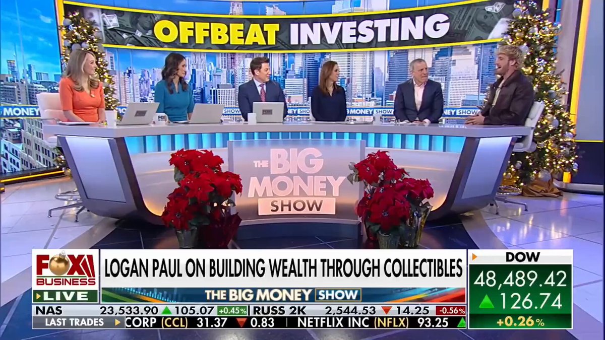 The image features a group of people sitting around a table, with a TV screen displaying a news segment about Offbeat Investing. The hosts are discussing the topic, while several guests can be seen seated at the table. In front of them is a potted plant and two vases filled with flowers, adding to the festive atmosphere. The overall scene suggests that this gathering could be taking place during the holiday season or around a special event where people have come together for a discussion on financial topics.
