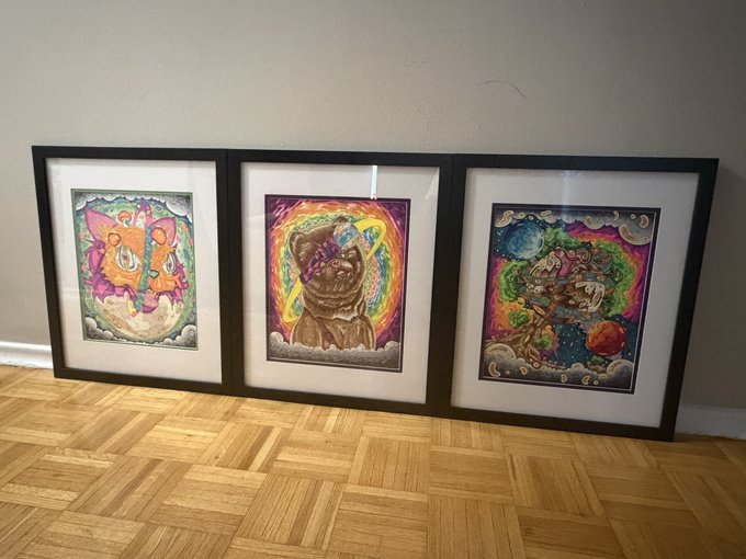 Three framed, vibrantly colored original artworks lean against a wall on a parquet floor, each featuring whimsical, psychedelic illustrations—including a cat in space and figures on clouds—showcasing the physical pieces that can be redeemed by buyers of the artist's 1/1 NFTs.