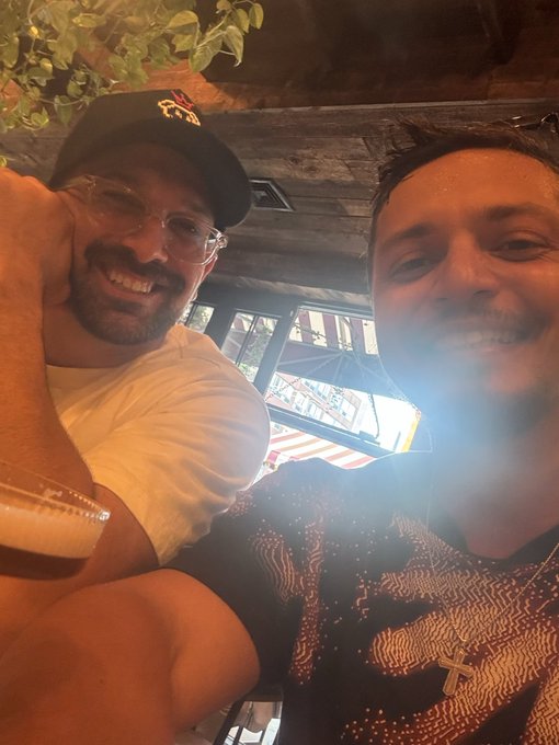 Two men smile for a selfie in what appears to be a restaurant or bar; the man on the left wears a cap and glasses, while the man on the right sports a mustache and a black shirt with an orange pattern.