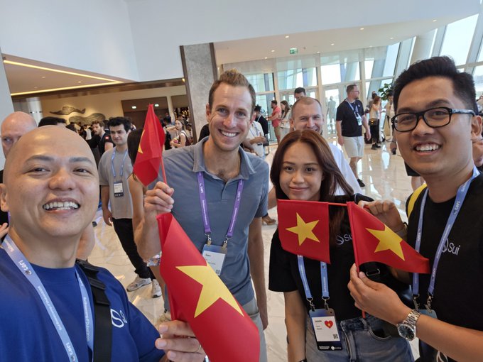 A group of smiling people pose for a photo at what appears to be a conference, with three of them holding small red flags featuring a single yellow star.