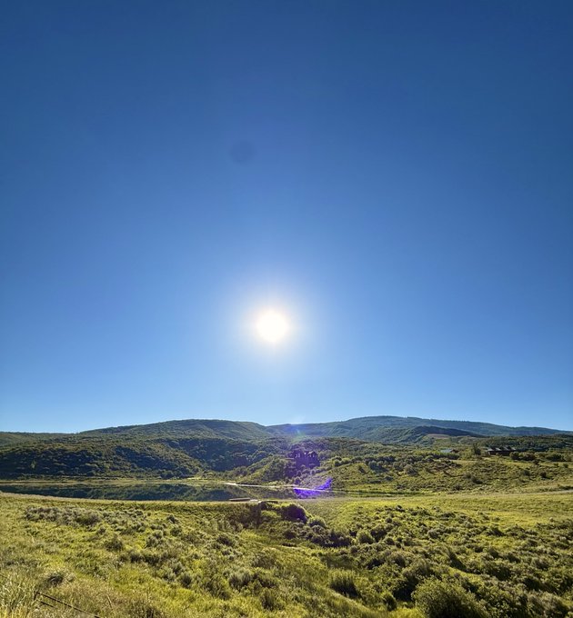 A bright sun shines high in a clear blue sky over a vast, rolling green landscape with distant mountains.