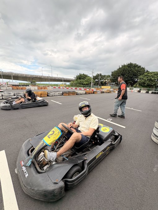 Two people wearing helmets sit in go-karts on an outdoor track, with a man in a red and white jacket walking nearby as an observer.