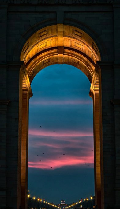Framed by a grand illuminated stone archway, a breathtaking sunset of pink and blue hues stretches across the sky with birds silhouetted in flight, leading down to a glowing pathway. The image's hopeful light at the end of a tunnel beautifully mirrors the caption's message of resilience, wishing for strength, recovery, and good health during a difficult month.