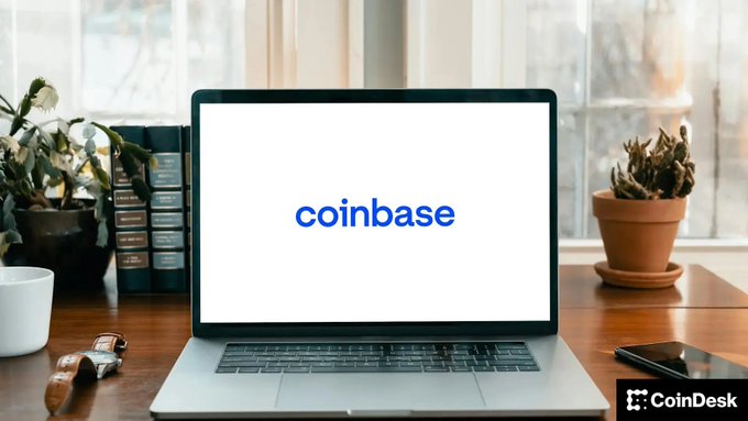 A laptop on a wooden desk displays the blue 'coinbase' logo, with a 'CoinDesk' watermark in the bottom right corner.
