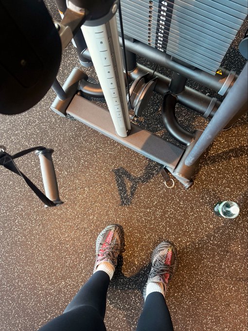 A first-person view from above looking down at a person's feet in colorful knit sneakers standing on a rubber gym floor in front of weight machines.