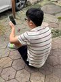 A small, low-resolution photo showing a person from behind wearing a striped shirt and a backpack, crouching on a tiled floor while using a smartphone.